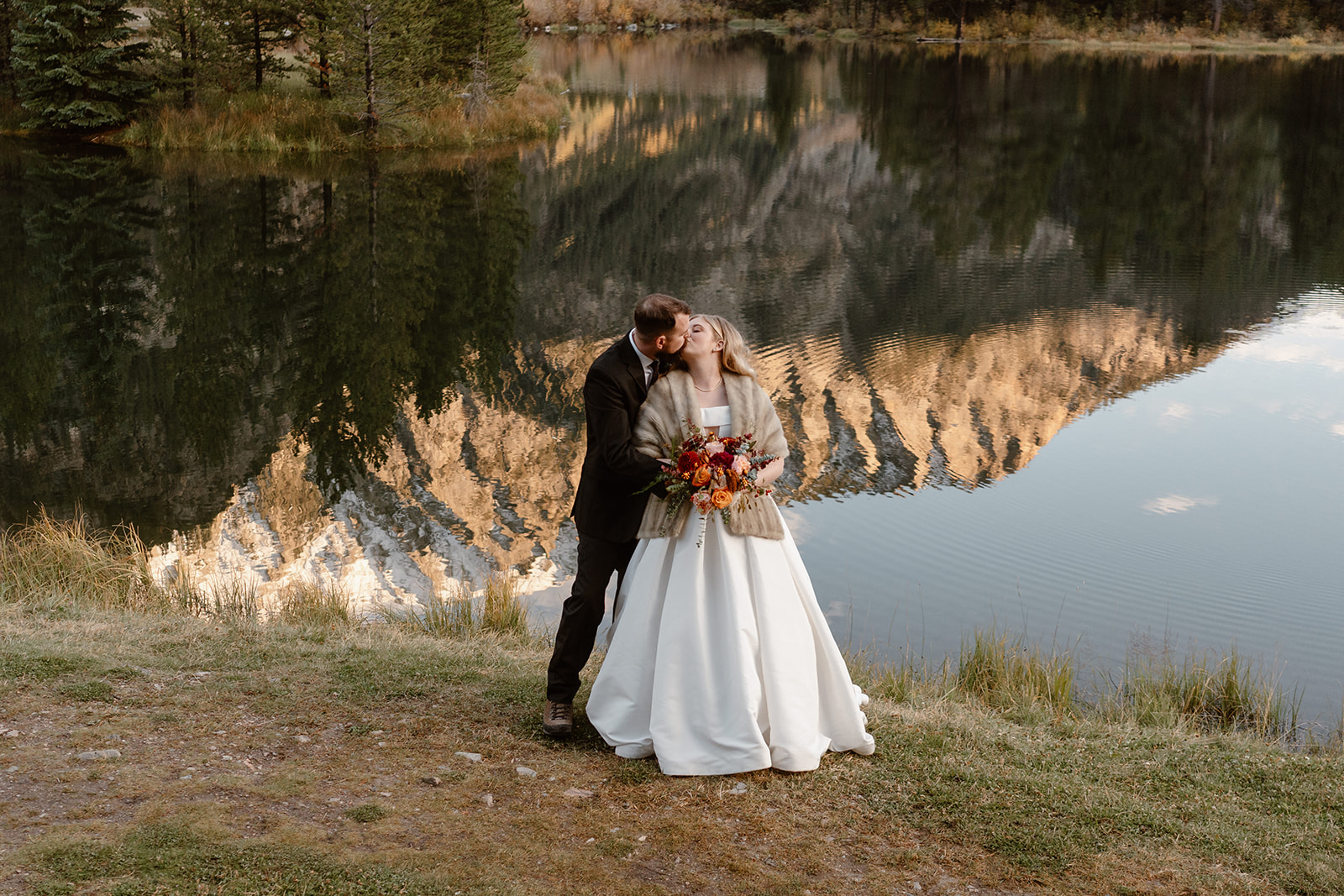 A bride and groom kiss in front of a reflective, Colorado alpine lake during their adventure elopement ceremony.