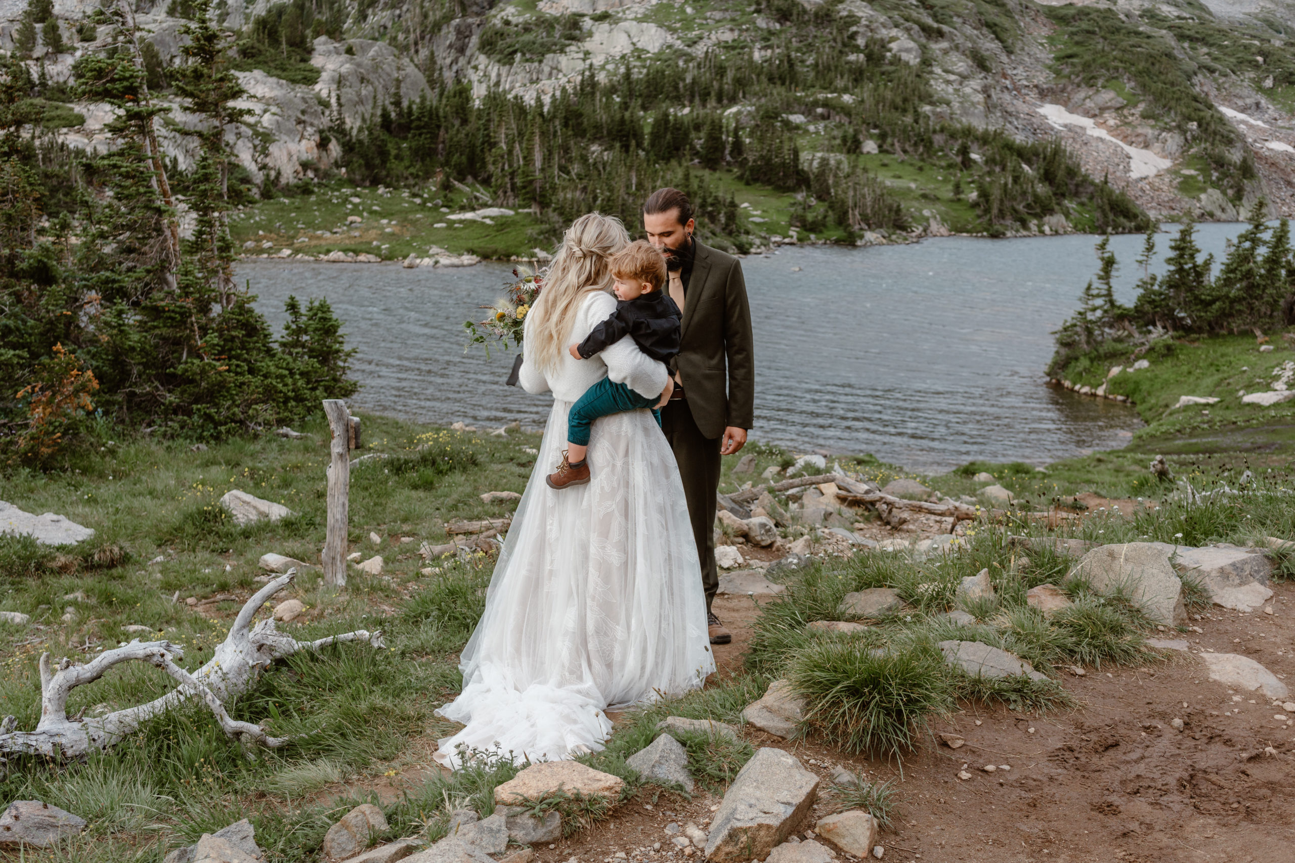 A bride holds her son as she approaches her soon-to-be husband near a lake in Colorado during their adventure elopement ceremony.