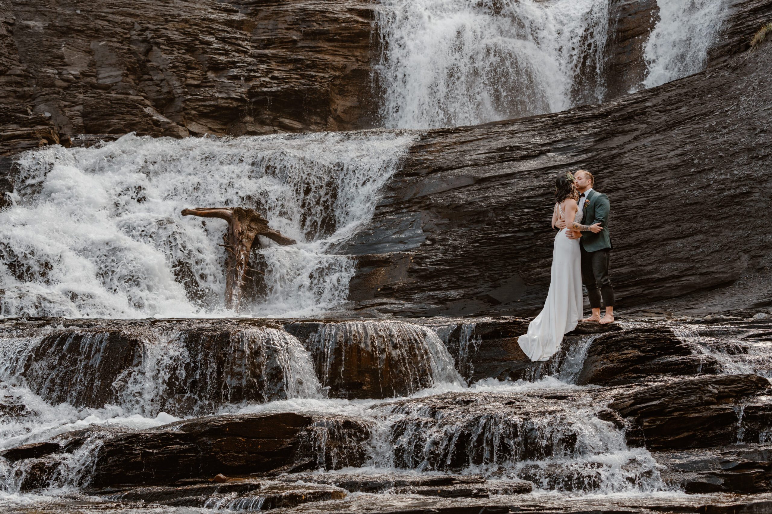 A bride and groom pose and kiss on top of a waterfall during their Colorado waterfall elopement. 