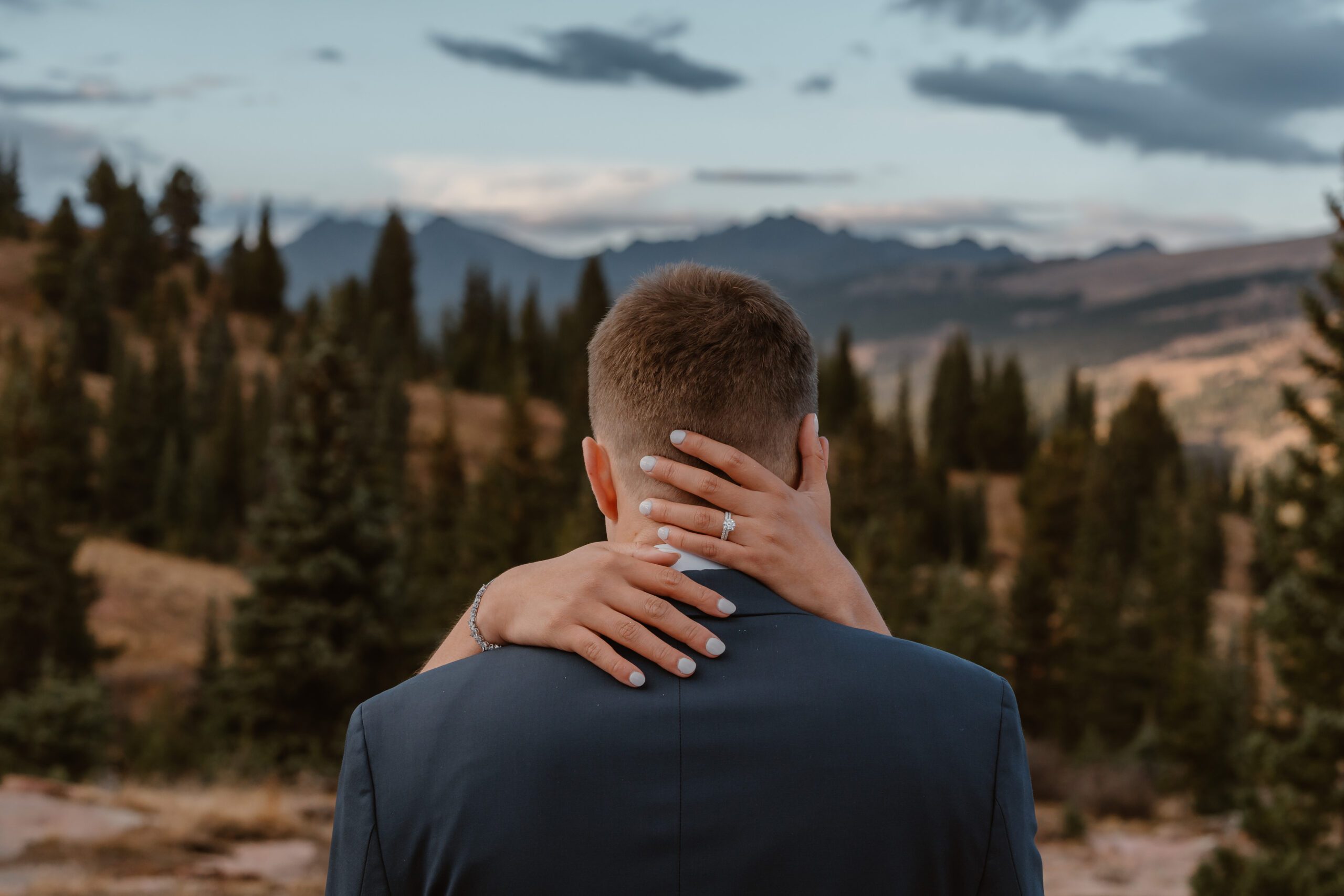 A groom is shown from the back with his bride's hands carefully wrapped around his head, her engagement ring and wedding band shown.