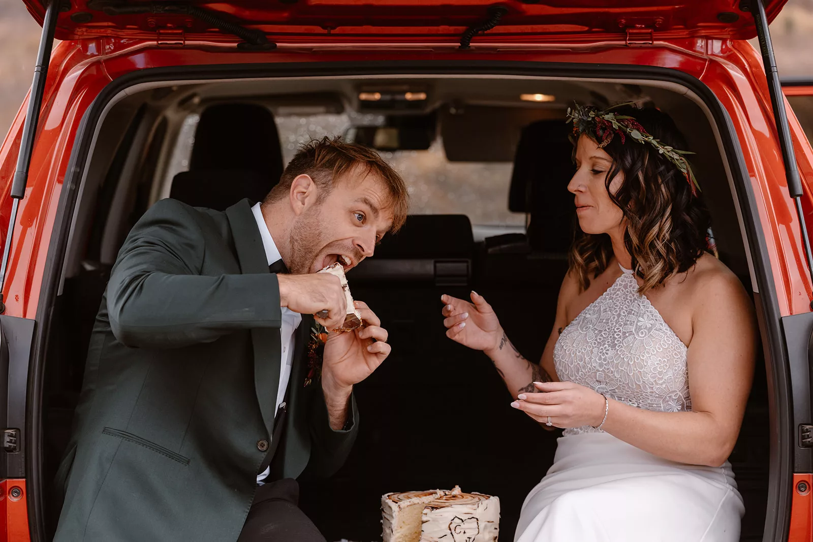 A bride and groom sit in the back of their truck eating their wedding cake before their adventure wedding hike.